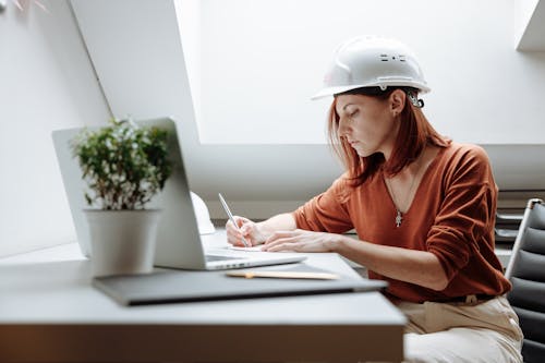 Woman Wearing White Hard Hat While Writing on a White Paper