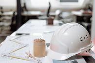办公桌特写，上面有白色头盔、彩色铅笔和建筑平面图