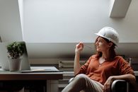 一位白人女建筑师头戴安全帽，坐在办公桌前，手持笔沉思