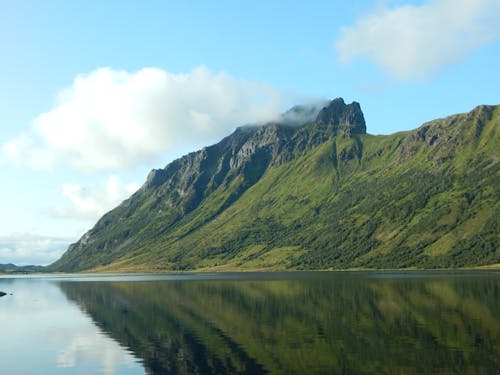 Majestic green slope of rocky range in clouds reflecting in tranquil water of clear lake