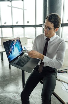 Young businessman analyzes data on a laptop in a high-rise office setting.