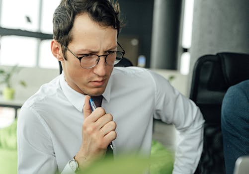 Man in white shirt thinking deeply at office desk with pen.