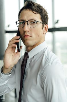 在明亮的办公室里，一个戴着眼镜的严肃男人正用智能手机交流
