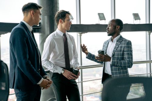Free Three businessmen in formal attire engaged in a conversation while holding coffee cups in a modern office setting. Stock Photo