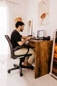 Side view of focused young man sitting at workplace with laptop and various gadgets and working at home in daytime