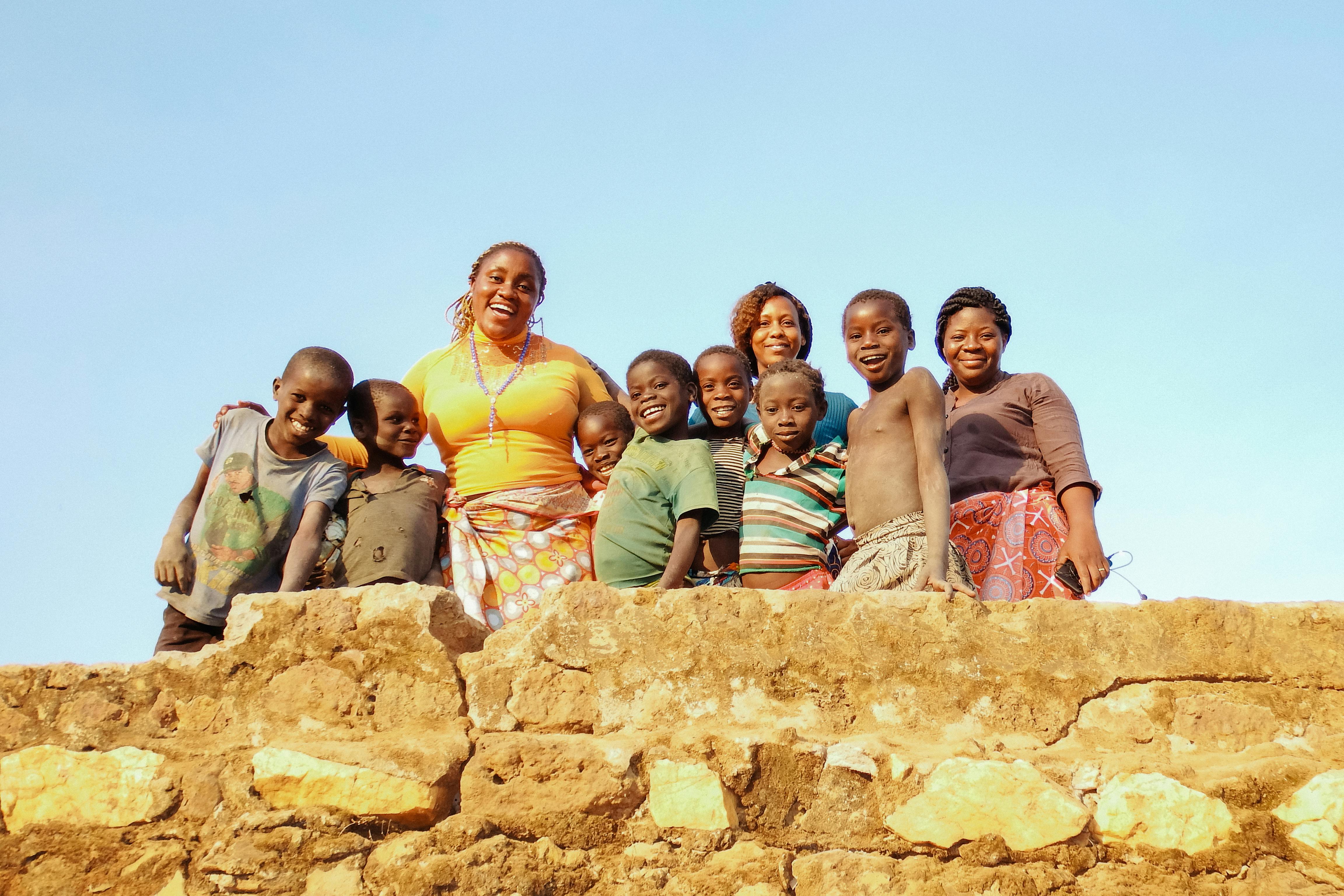 A cheerful group of African children and women standing behind a stone wall under a clear sky.