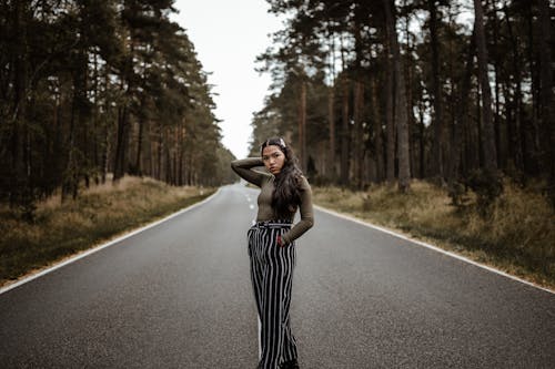 Calm young ethnic female in trendy wear standing on empty roadway between lush trees while touching head and looking at camera