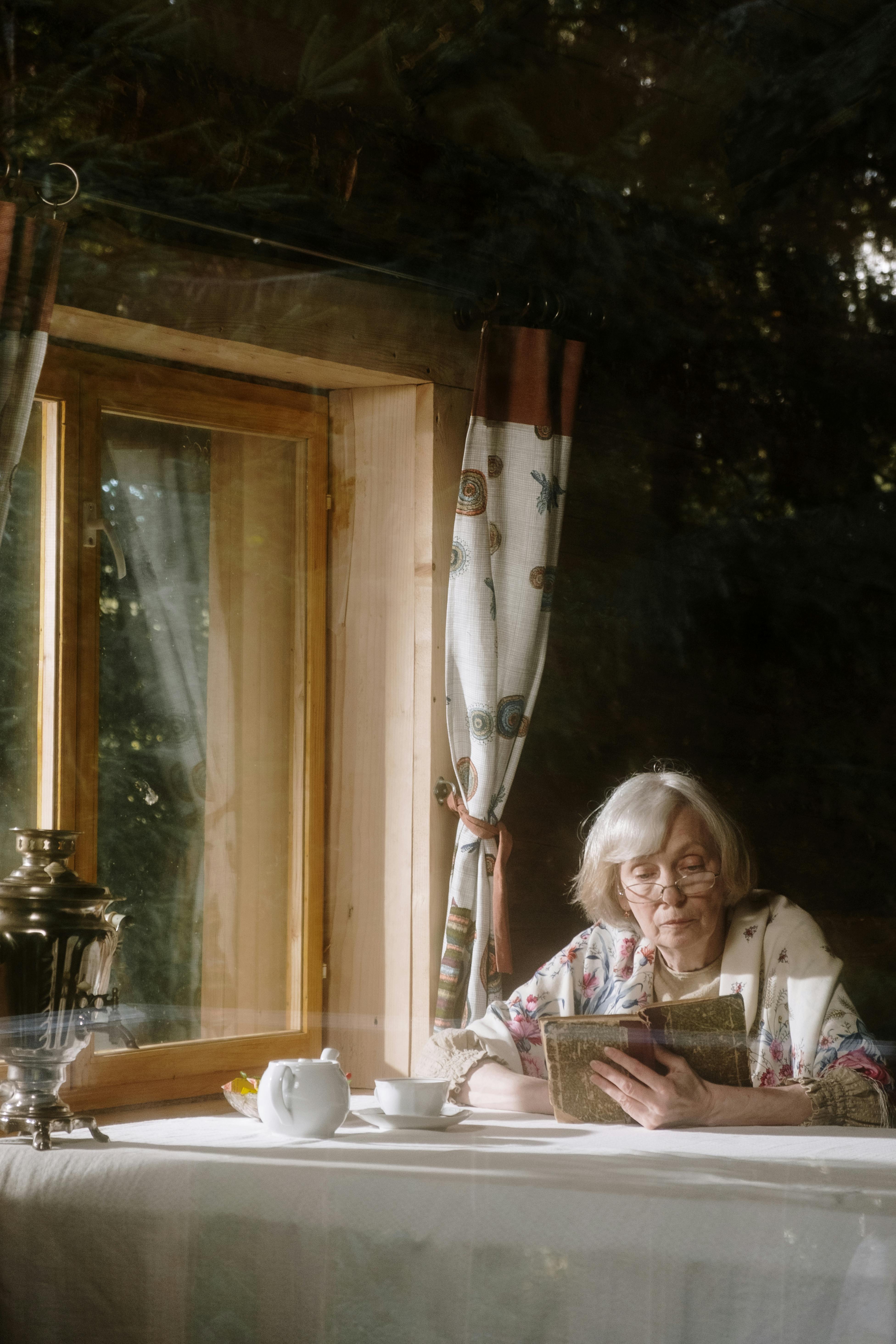 elderly woman reading a book beside a window