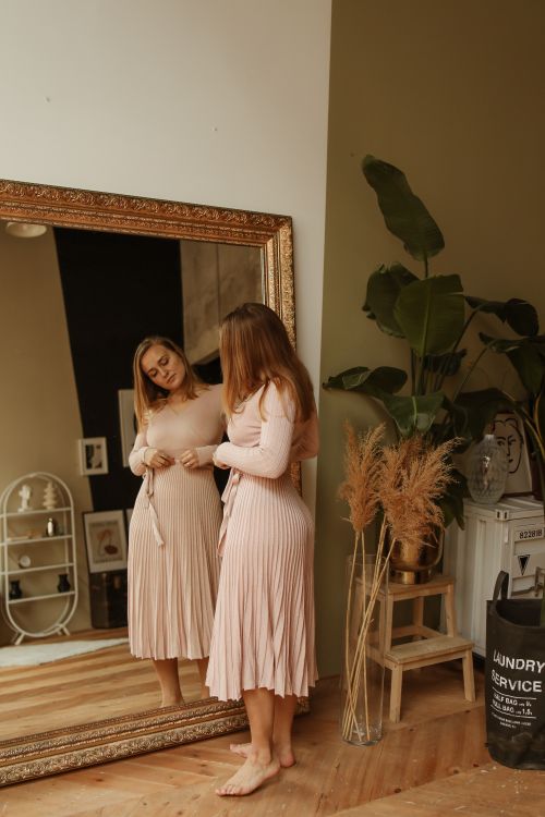 Free Adult woman in pink dress adjusts outfit in a stylish room with art and plants, examining her reflection in a large mirror. Stock Photo