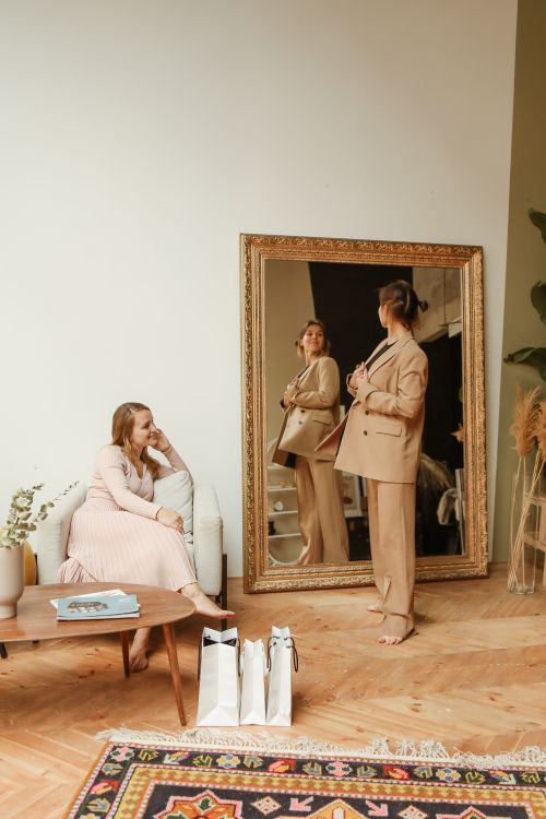 Free A woman in a suit admires her reflection in a large ornate mirror, with another woman seated nearby. Stock Photo