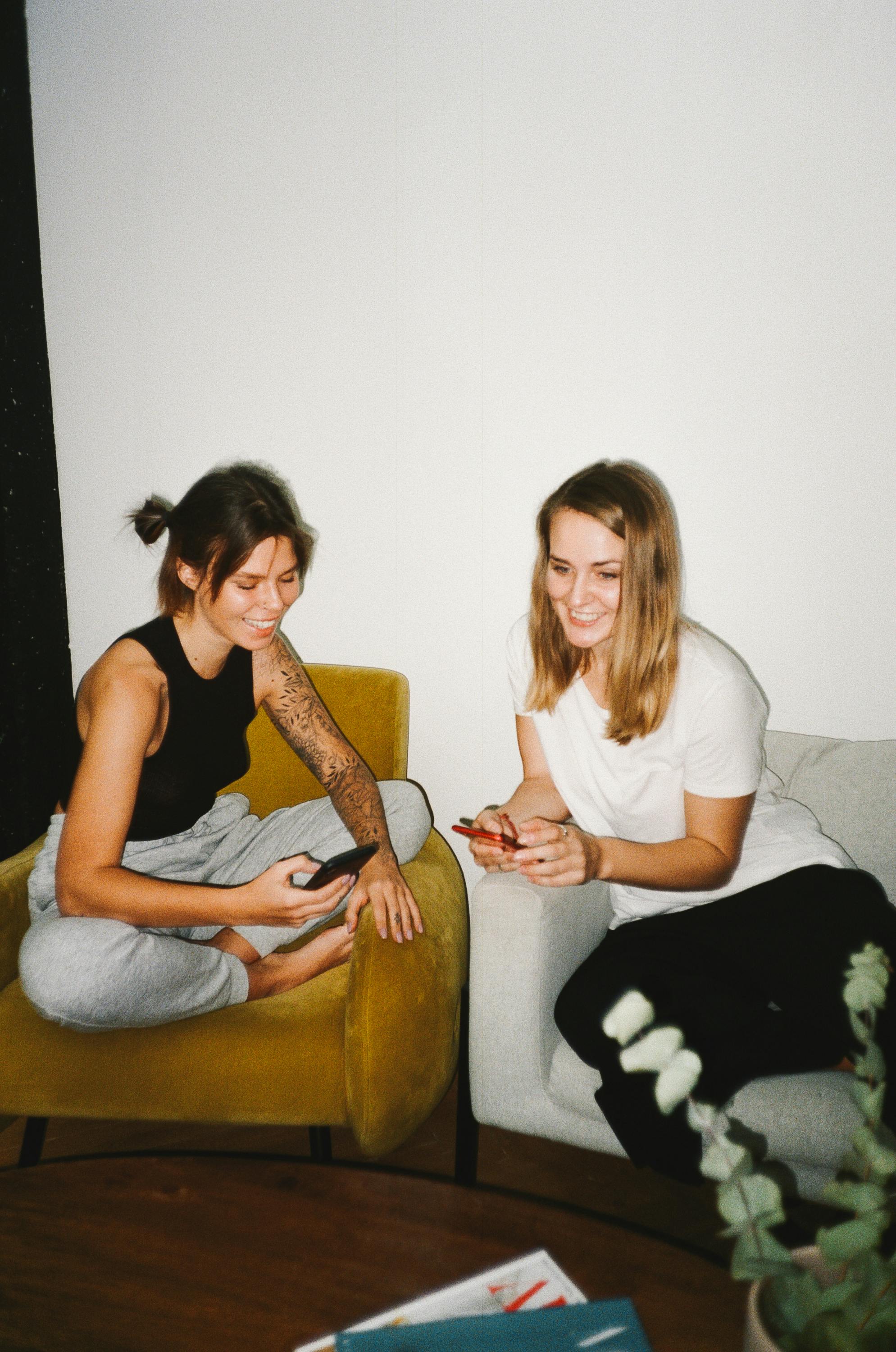 Two women smiling and using smartphones indoors, sharing a fun moment.