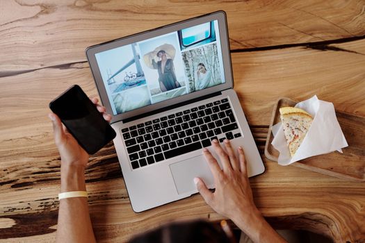 Person uses smartphone and laptop while enjoying a sandwich at a wooden table, showcasing multitasking and technology use.