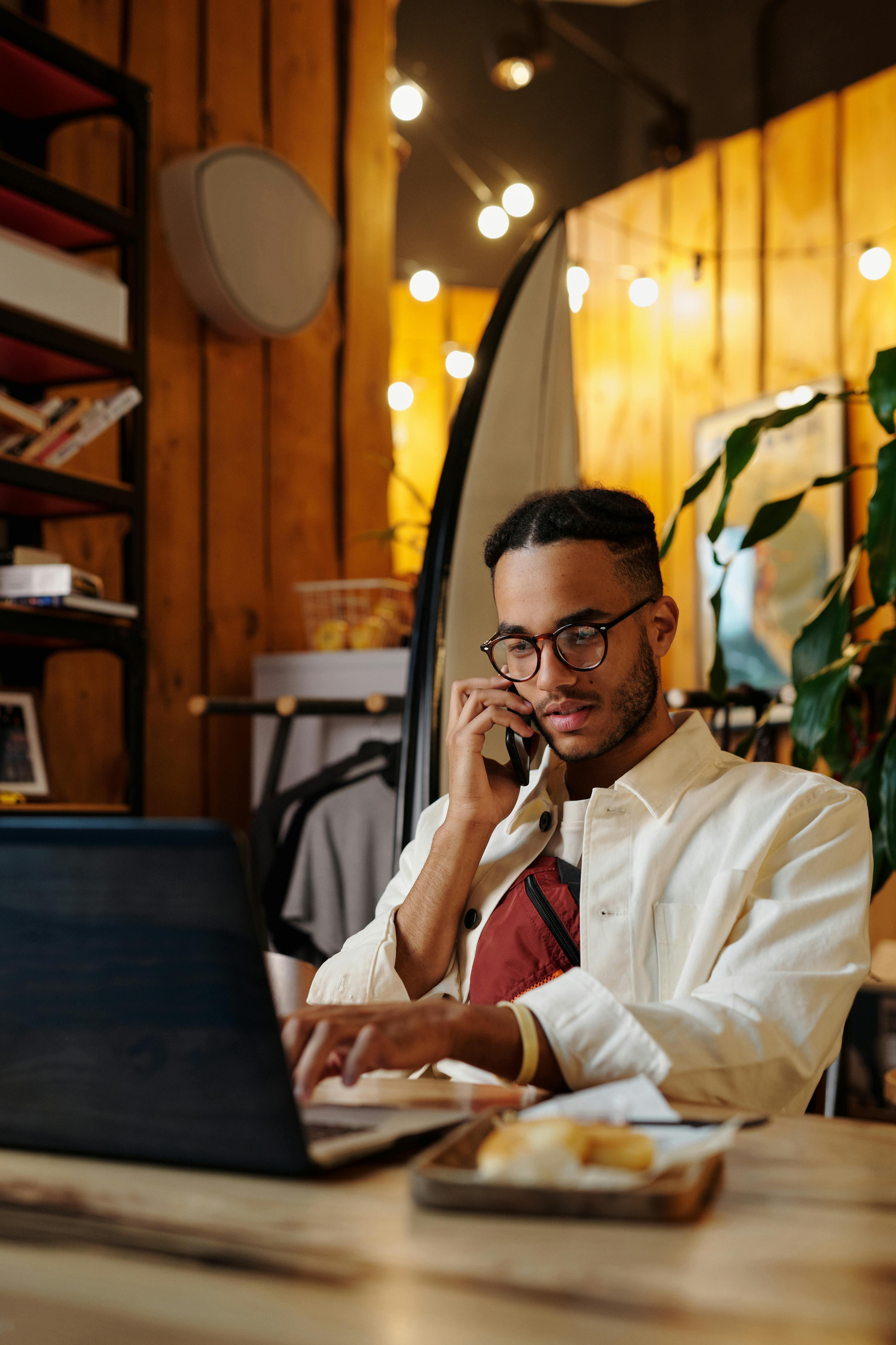 A stylish freelancer multitasking on phone and laptop in a cozy, well-lit café.