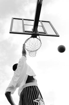 Low-angle black and white shot of a man dunking a basketball outdoors.