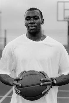 Monochrome portrait of a male athlete holding a basketball on an outdoor court.