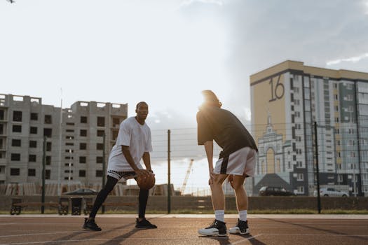 Two young men playing basketball on an outdoor court during sunset near tall buildings.