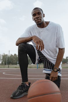 A man kneeling on an outdoor basketball court, wearing activewear, posing confidently during the day.