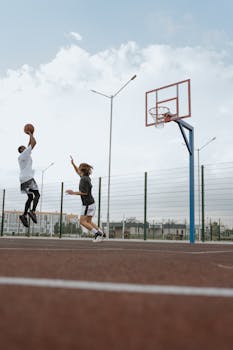 Two young men playing basketball on an outdoor court with one shooting midair.