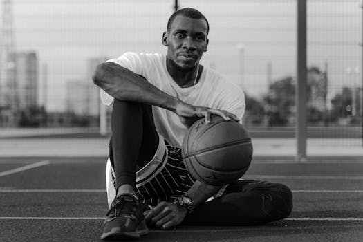 Monochrome image of a man sitting on a basketball court holding a ball.