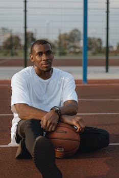 A young man sitting on an outdoor basketball court holding a basketball, expressing relaxation and sportsmanship.