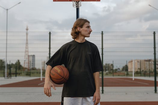 Profile of a young man holding a basketball on an outdoor court under a cloudy sky.