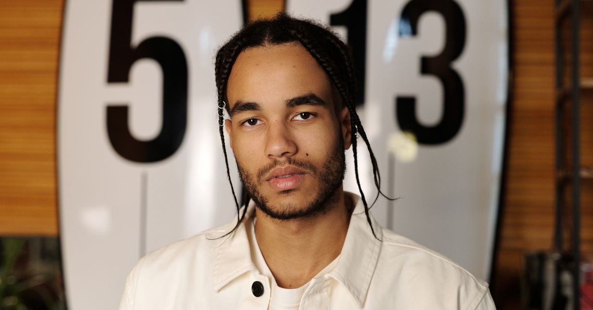 A stylish man with braids poses in front of surfboards marked 5 and 13.
