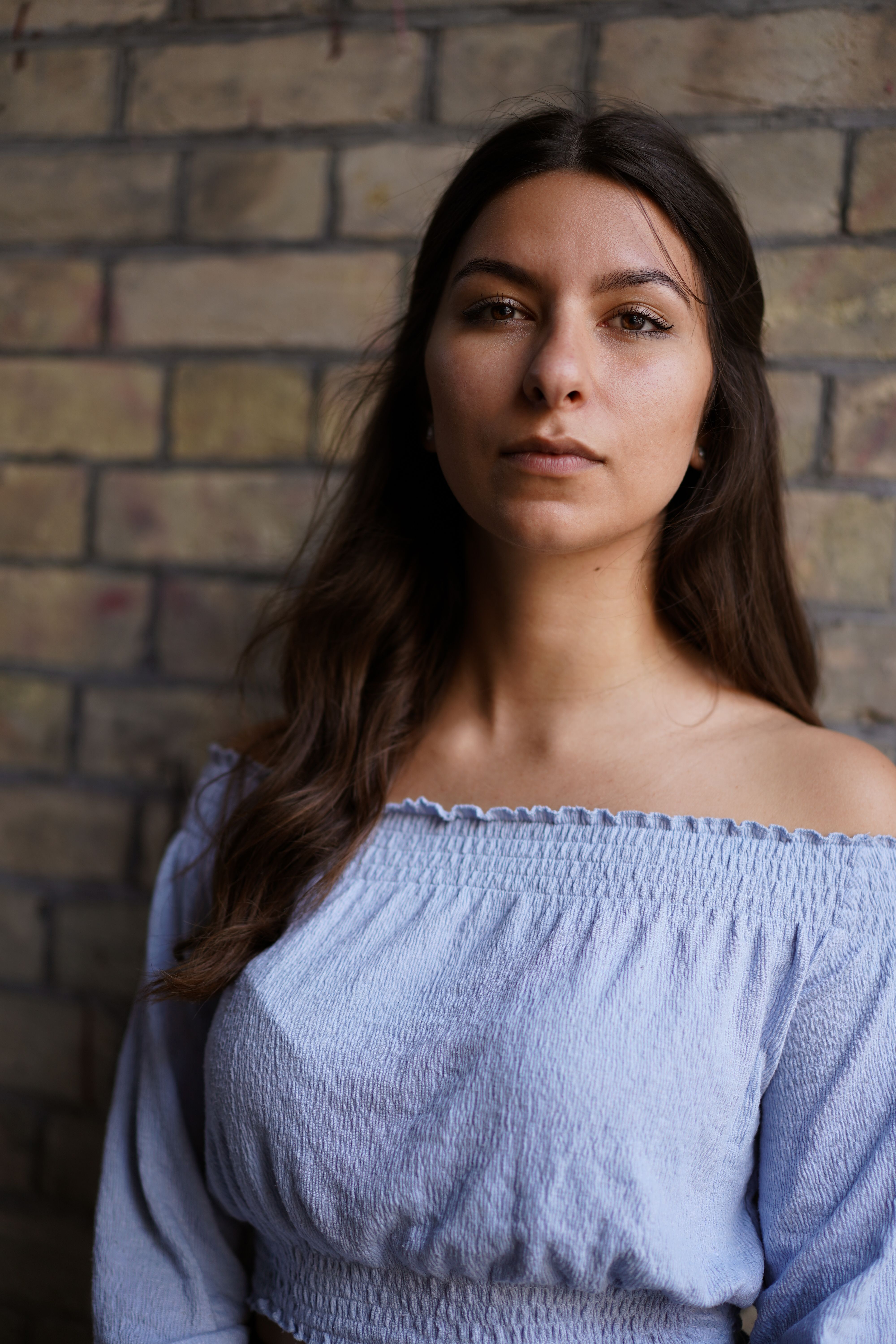 Portrait of a young woman in a blue off-shoulder top against a brick wall, exuding elegance and confidence.