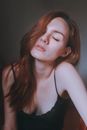 Beautiful female with tilted head and closed eyes wearing black top with thin straps sitting in room against gray background