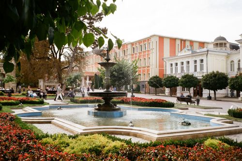 Free Fountain in park with bushes and trees near street of modern town with classic buildings Stock Photo