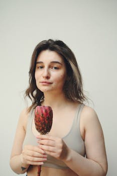 Young woman in sports bra poses with a flower against a neutral background, embracing natural beauty.