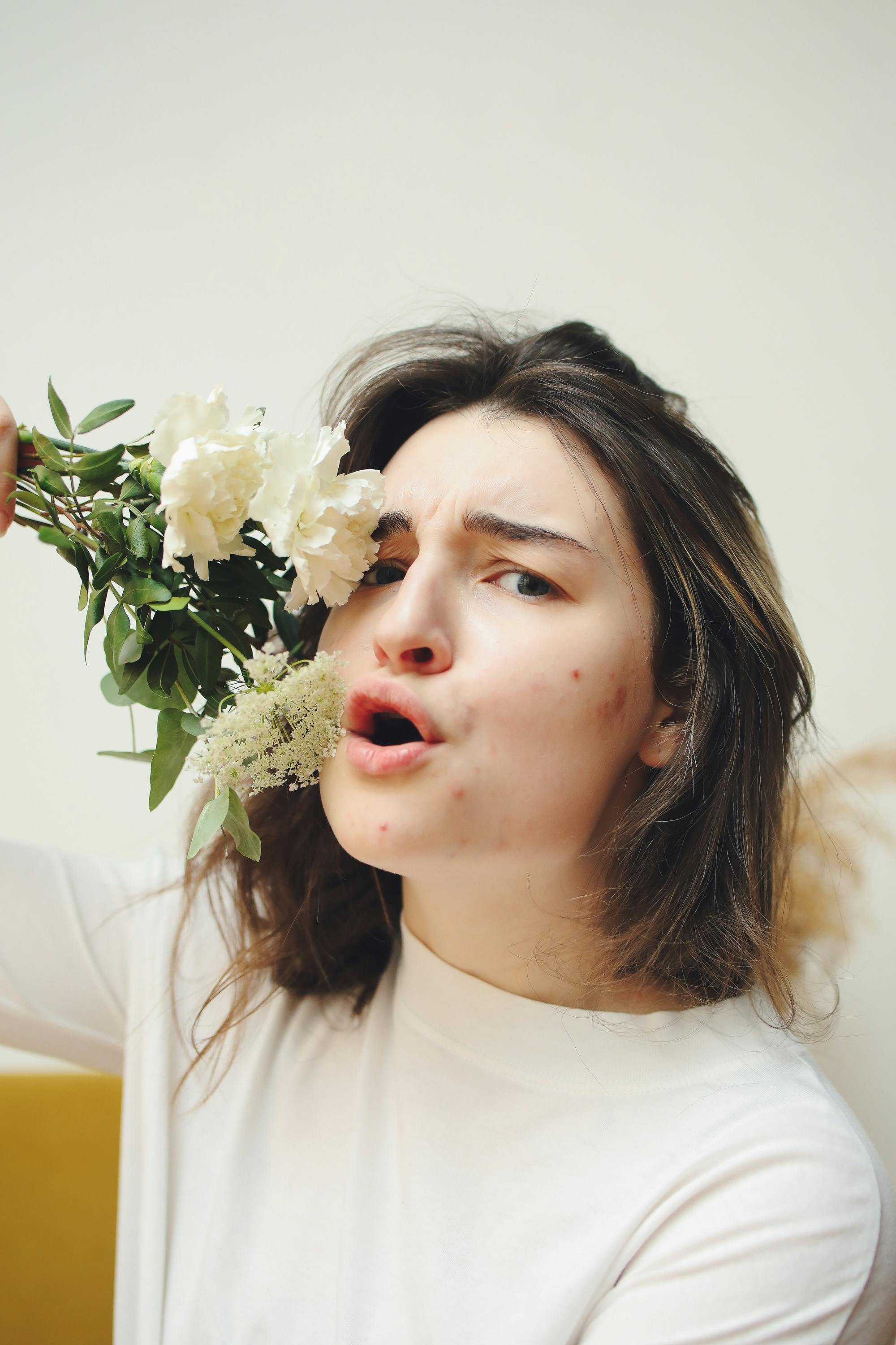 A woman with acne speaks expressively while holding white flowers, with a neutral background.