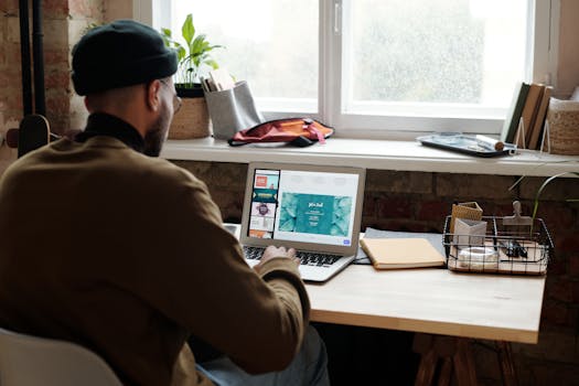 A man working remotely at home, focusing on a laptop while seated at a desk by a window.