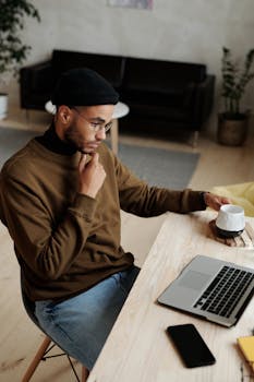 A young man in a casual workspace thinking while working remotely on his laptop with a cup of coffee.