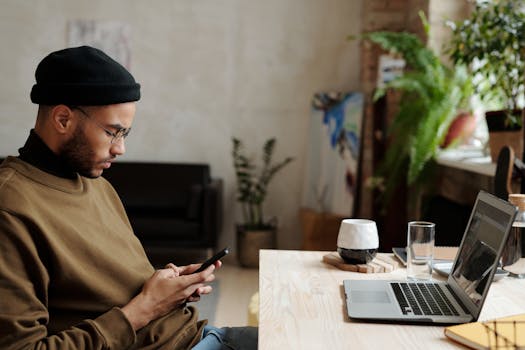 Young man using his phone at a serene home office with a laptop nearby, embodying remote work lifestyle.