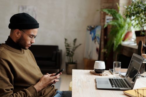 Free Young man using his phone at a serene home office with a laptop nearby, embodying remote work lifestyle. Stock Photo