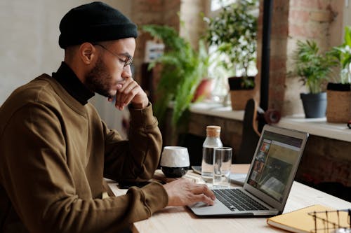 мужчина в коричневом пальто и синей вязаной шапке сидит за столом с Macbook Pro