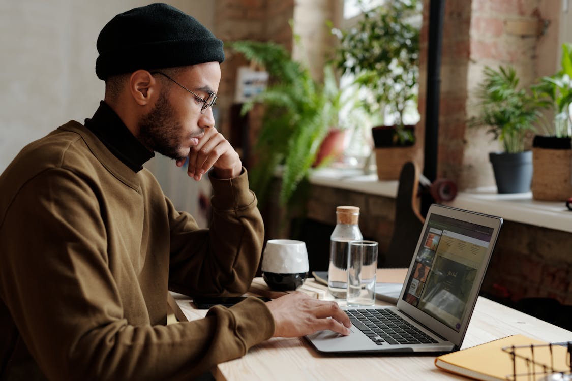 Foto Hombre con abrigo marrÃ³n y gorro de punto azul sentado junto a la mesa con Macbook Pro de stock gratuita