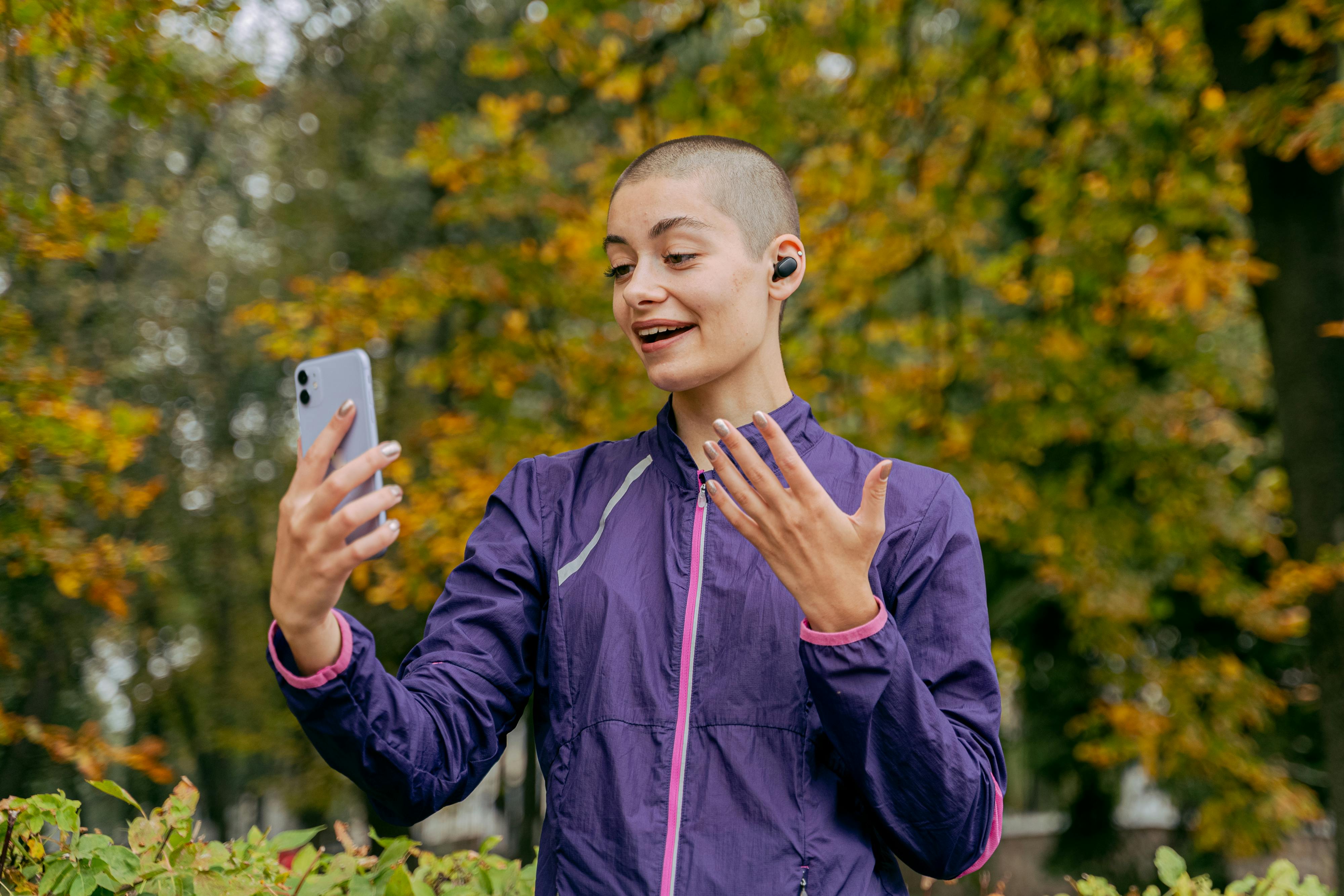 A woman in active wear makes a video call outdoors during autumn, surrounded by colorful trees.
