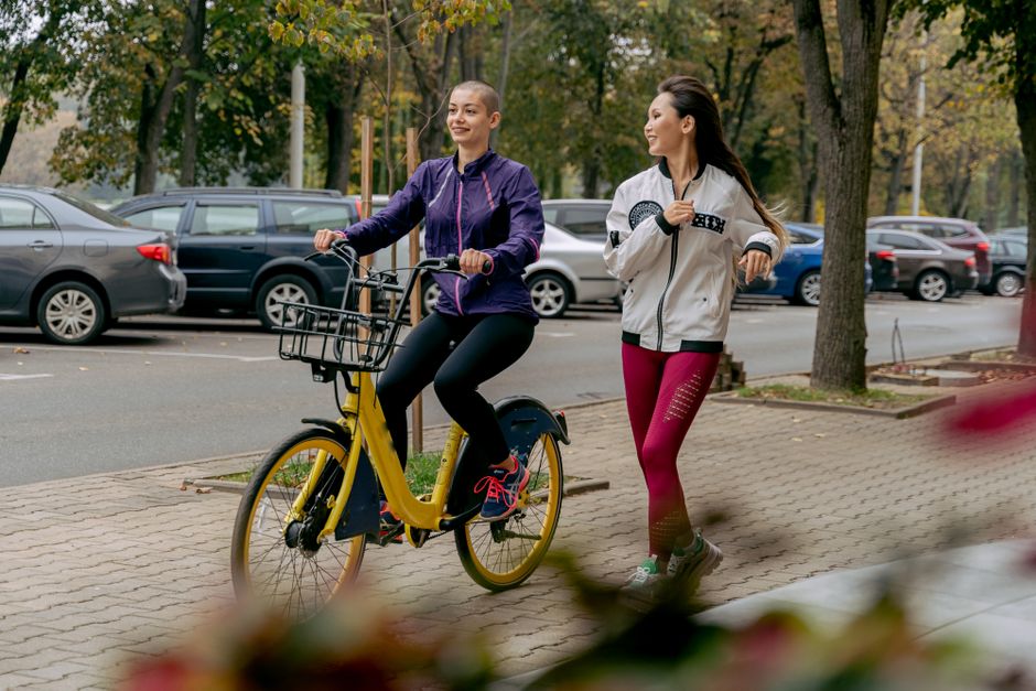 Two women enjoying an outdoor exercise session, cycling and jogging on a city sidewalk.