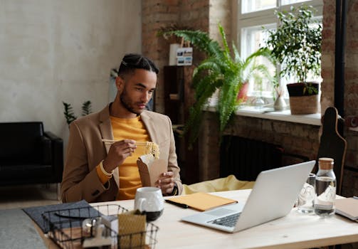 A young man enjoying takeout noodles while working at a modern office desk.