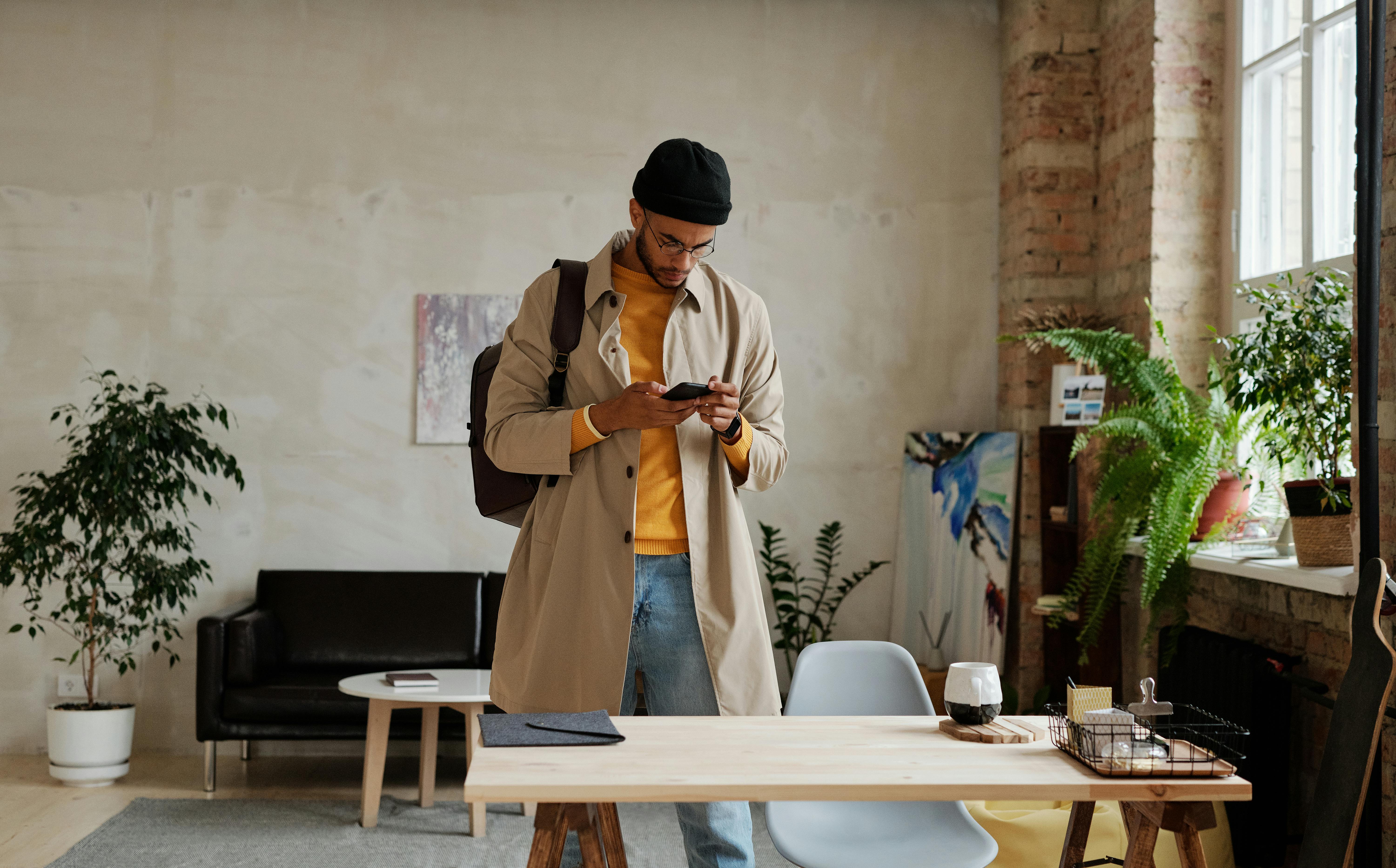 一位穿着米色外套的时尚男子，在摆满植物和艺术品的别致城市阁楼里查看手机