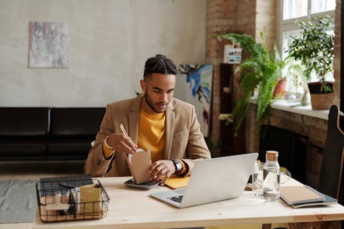 Free Man in Brown Blazer looking at a Laptop while eating Noodles Stock Photo