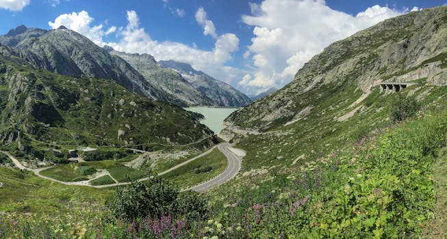 Panoramic view of green mountains, a winding road, and a serene lake in Obergoms, Switzerland.