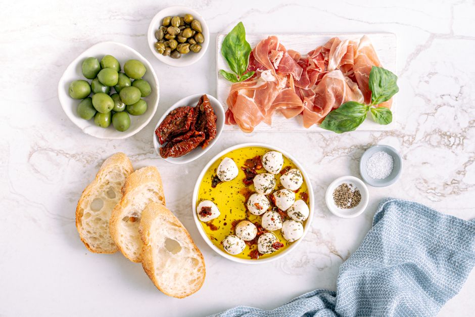 A wooden board arranged with sliced prosciutto, marinated olives, cubes of parmesan, bruschetta topped with tomato and basil, and a small bowl of balsamic glaze.