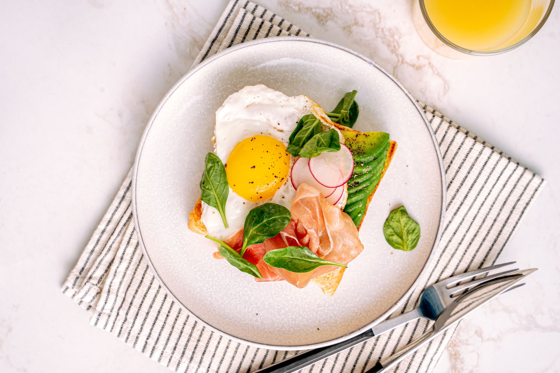 A mouth-watering breakfast toast featuring a fried egg, avocado, and prosciutto on a white plate.