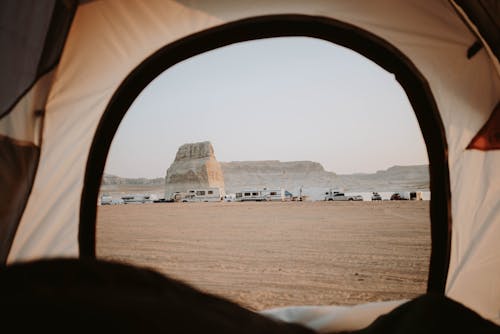 View of sandy beach with distanced cars parked on seaside with rocky cliffs through opened tent in nature with cloudless sky