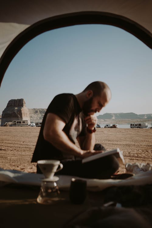 Side view of focused male tourist sitting near doorway of tent with filtered coffee and reading book in countryside during trip