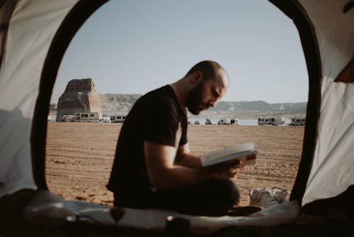 Side view of bearded male traveler sitting in tent with opened door and reading book in nature with parked cars in distance