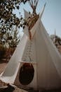 Traditional white cone shaped wigwam made of fabric with branches located in nature near tree sprigs with foliage in countryside
