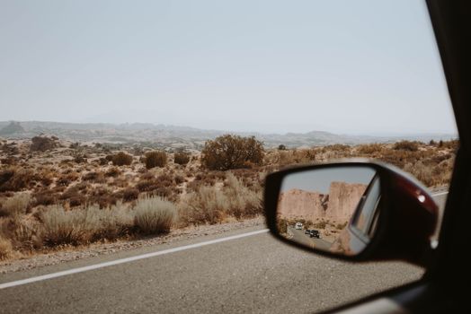 View of dry terrain with green shrubs through window of car with side view mirror riding on asphalt highway in nature
